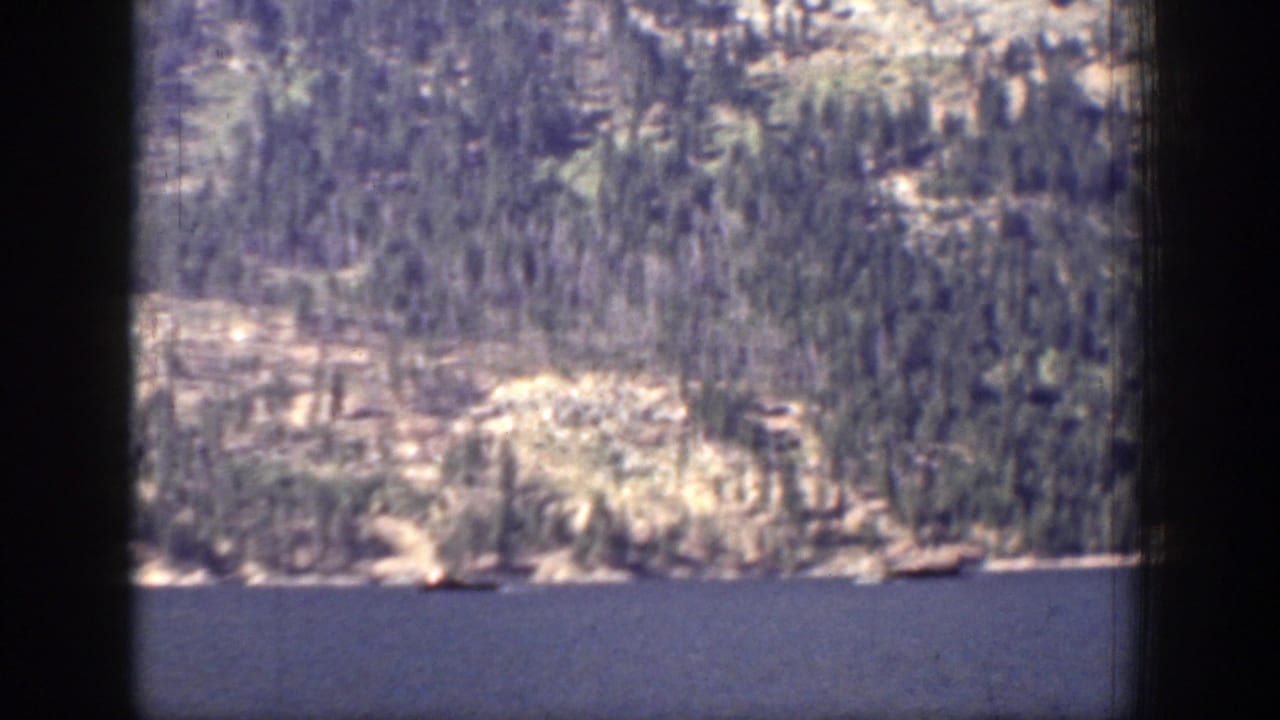 Wide lake with a small boat below forested mountain slopes and dark film borders.