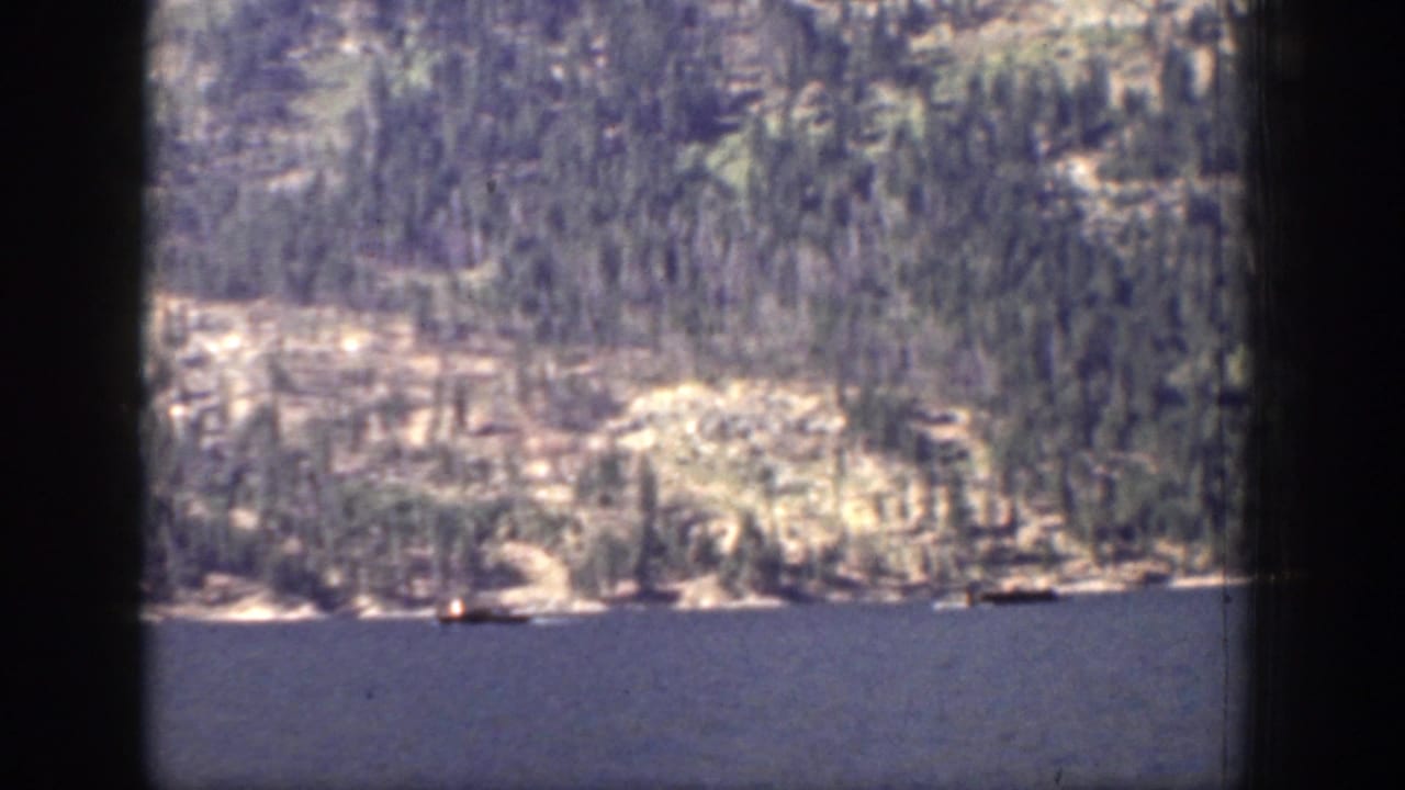 Chelan home-movie frame from 1939 showing a small boat on open water below a pale rocky shoreline and wooded slope.