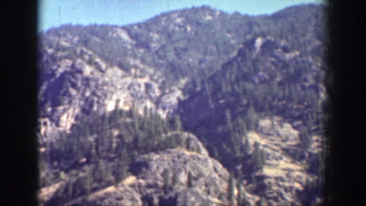Rocky forested mountainside under pale blue sky with dark film borders.