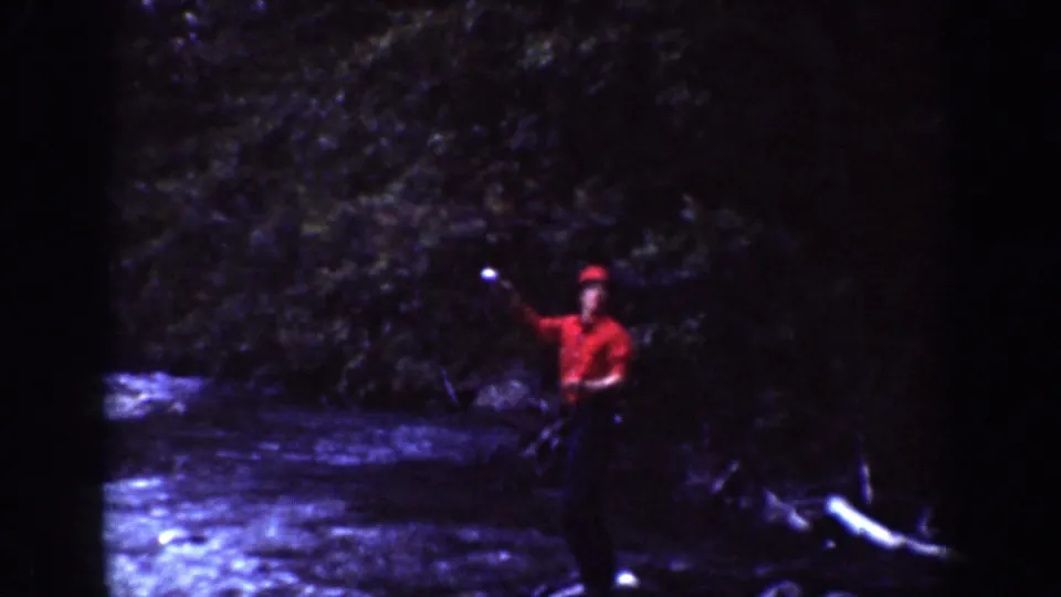 1939 Chelan, Washington home-movie frame showing a red-shirted adult by a river with dense forest behind.