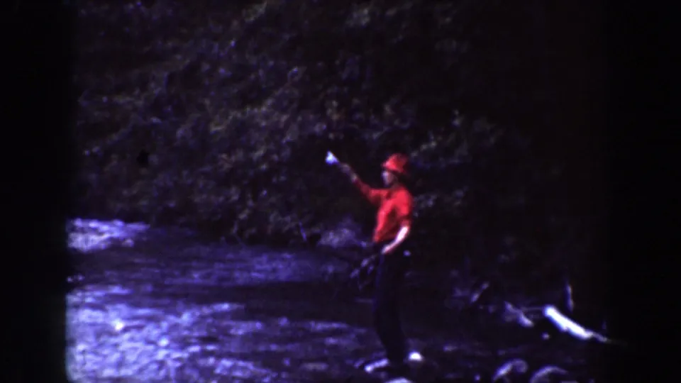 1939 Chelan, Washington frame showing a red-shirted adult near rushing water, with wooded slopes behind.