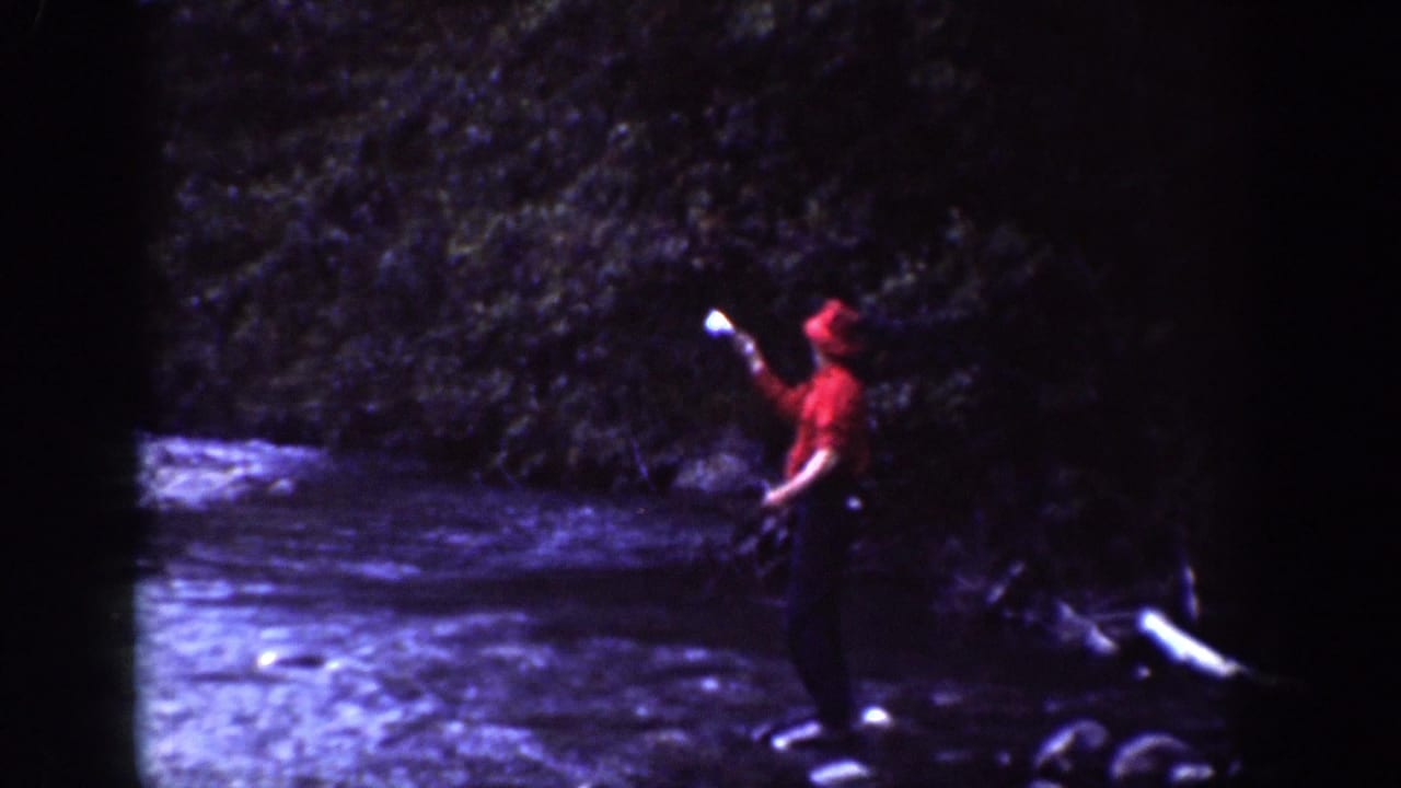 Man in red clothing stands in a shallow river holding a pale object near trees.