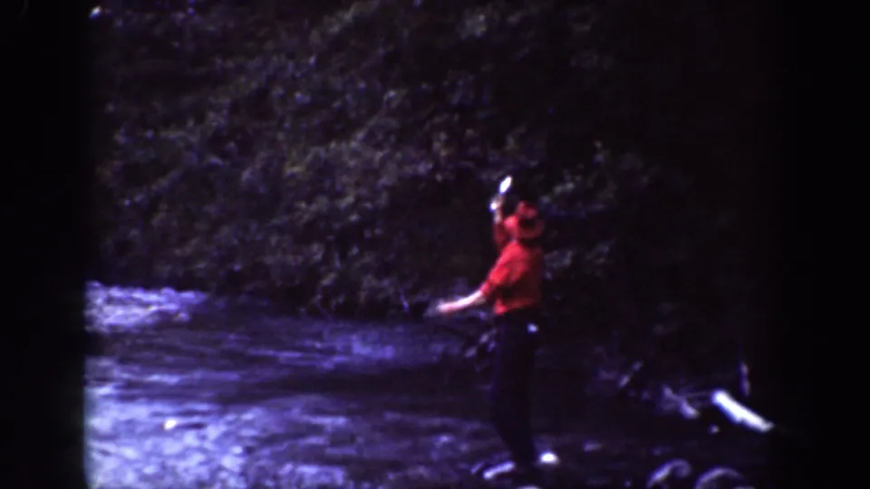 A man in red clothing stands at a wooded riverbank in Chelan, Washington, raising one arm while handling a fish or object.