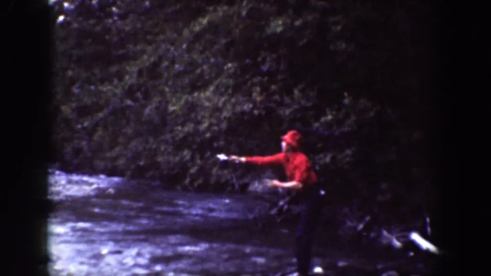 The man extends his arm over the shallow river, with trees and bank vegetation behind him in the 1939 Chelan home movie.