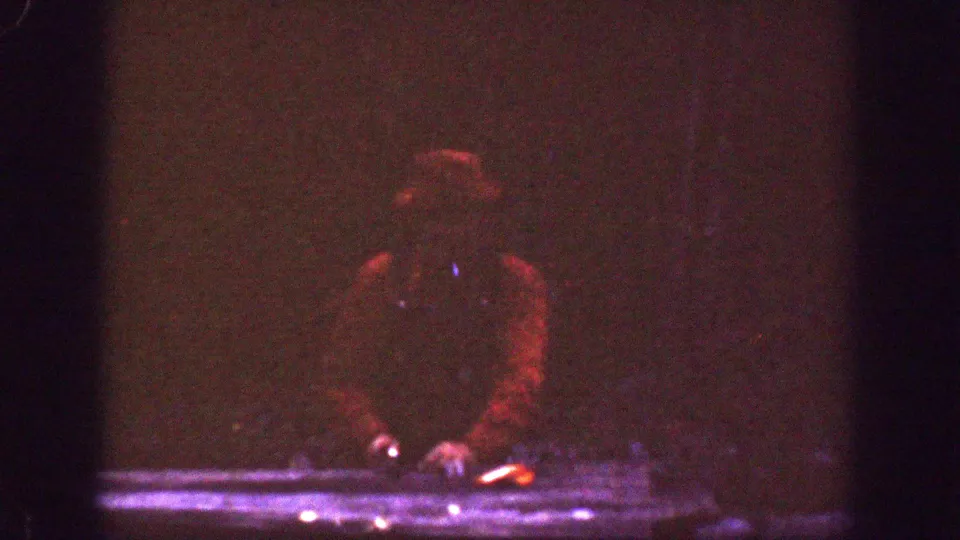 A man in dark clothing and a red cap leans over a table, with a small orange glow visible beside his hands.