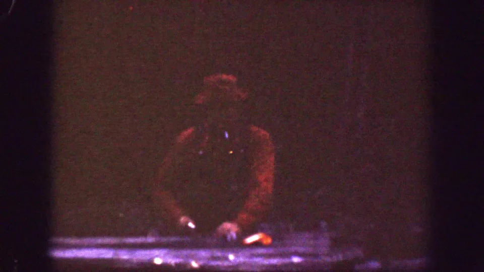 The man remains near the table in a very underexposed 8mm frame, with his cap and hand position faintly visible.