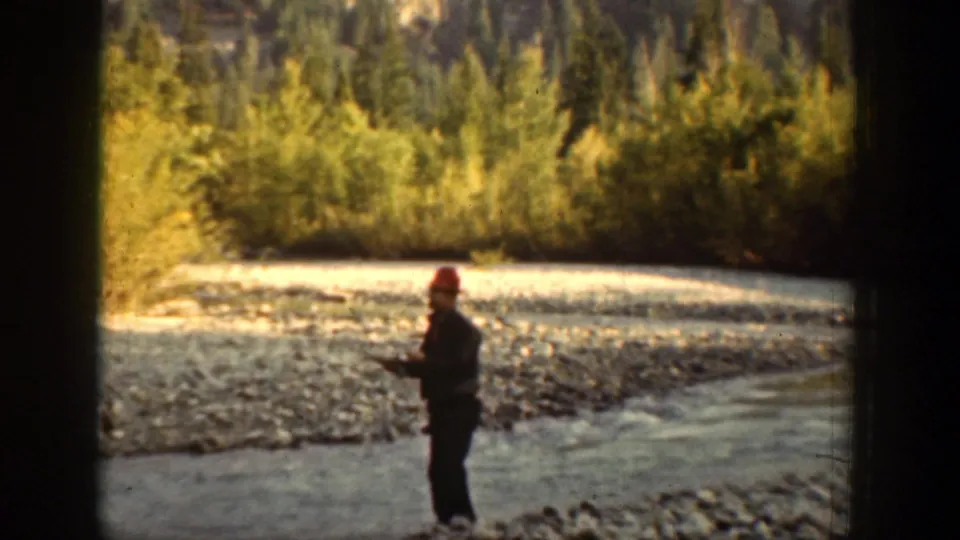 In Chelan, Washington footage, a man in a red cap stands in shallow river water, appearing to fish near a wooded bank.