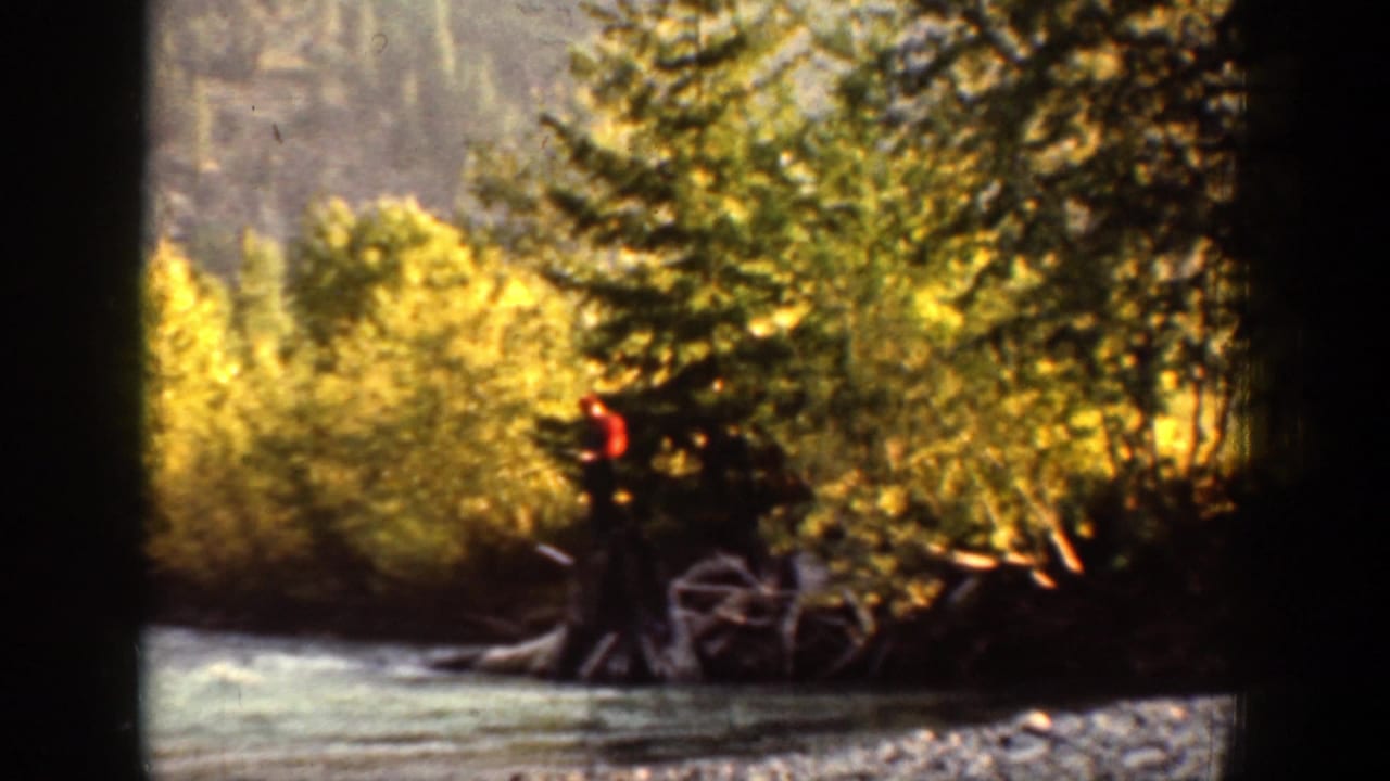1939 Chelan, Washington home-movie frame showing a distant man in red near a wooded riverbank and flowing water.