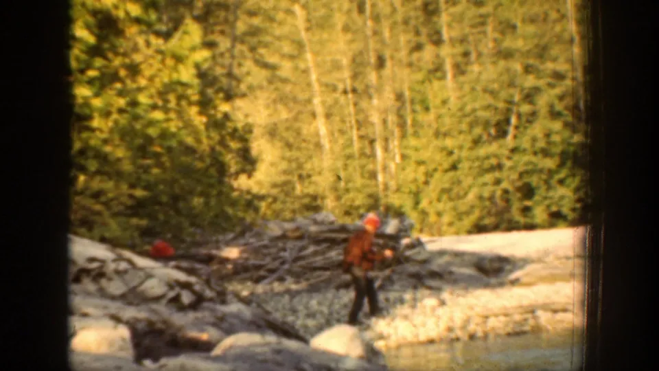 A man walks along a rocky riverbank in the Chelan, Washington home-movie scene, with forest and pale shoreline behind him.