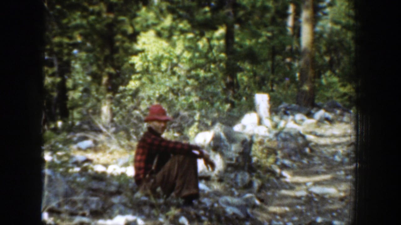 Man in a red hat and plaid shirt sits on a rocky forest trail.