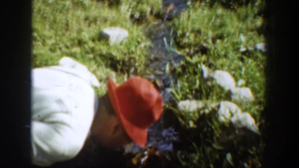 The man in the red hat leans down near a narrow stream bordered by grass and pale rocks in the 1939 Chelan footage.