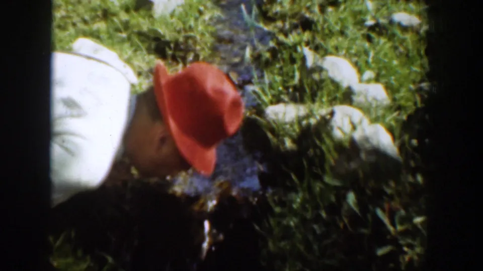 A close, soft 8mm view shows the man in a red hat leaning toward running water among grass and rocks in Chelan, Washington.