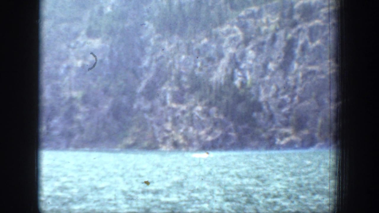 Small boat crossing a lake below a steep mountain shoreline, framed by dark film edges.