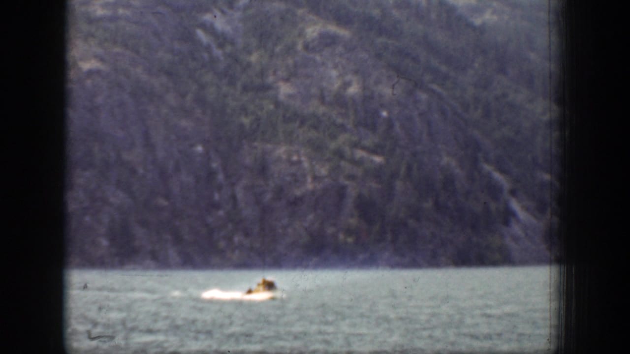 The motorboat continues across the Chelan lake scene, framed by dark vertical foreground edges and a steep wooded shoreline.