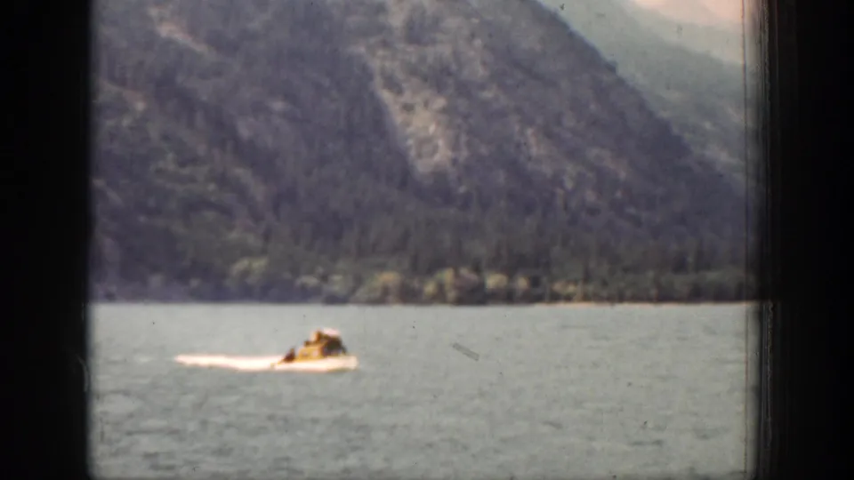 A small white motorboat moves across Lake Chelan, Washington, with forested mountains in the background in 1939 8mm footage.
