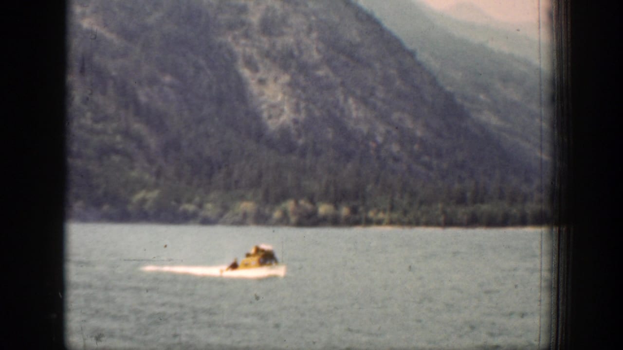 The motorboat continues across the Lake Chelan water, seen through soft 8mm grain with mountains and shoreline behind it.