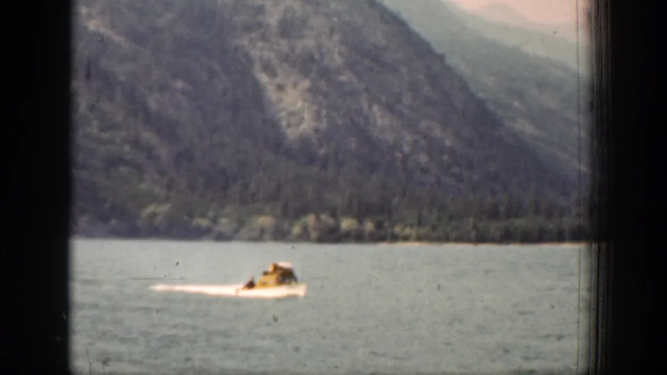 The motorboat continues across the Lake Chelan water, seen through soft 8mm grain with mountains and shoreline behind it.