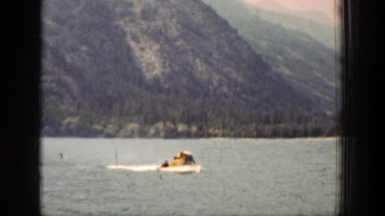 A small white boat with a visible rider passes along Lake Chelan, framed by water, shoreline trees, and hazy mountains.