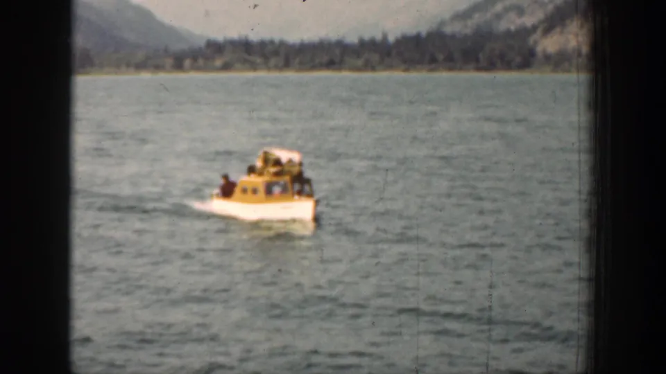 1939 Lake Chelan home-movie frame showing a small yellow-and-white boat moving across open water with forested hills in the distance.