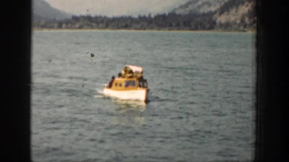 The yellow-and-white passenger boat continues over Lake Chelan water, with distant trees, mountains, and 8mm frame edges visible.