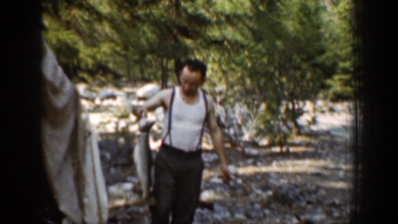 Man in suspenders walks on a rocky riverbank holding a large fish.
