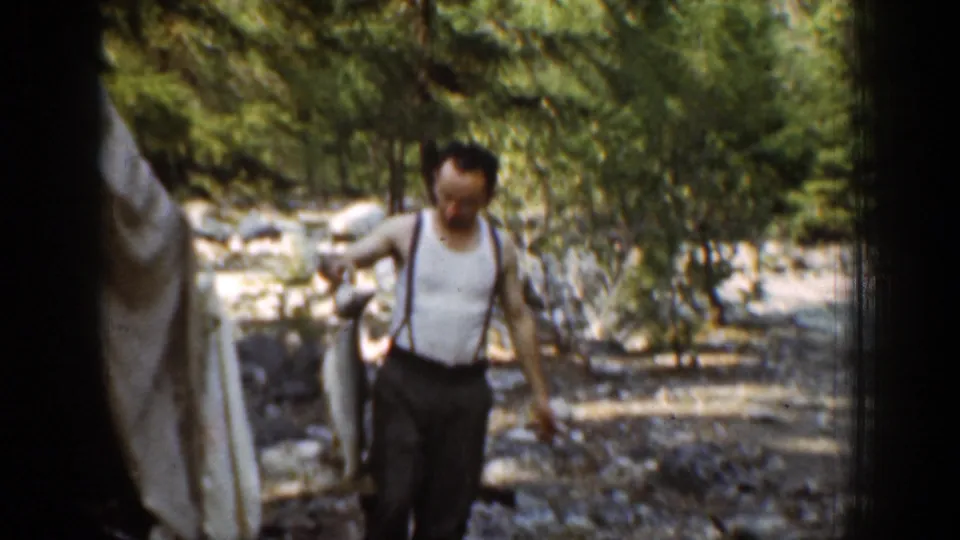 In Chelan, Washington home-movie footage from 1939, a man in a white tank top and suspenders carries a large fish along a rocky stream bank.