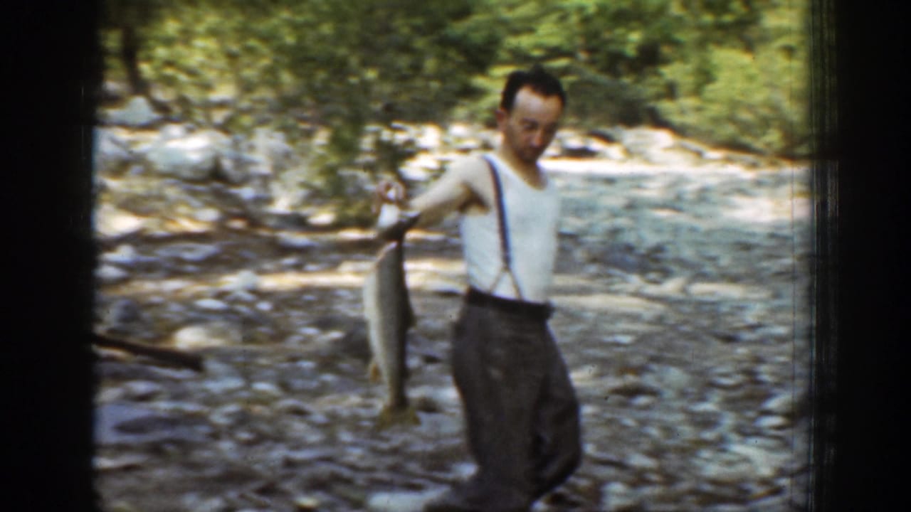 The man stands on a rocky stream bed in Chelan, Washington, holding a large grey fish beside him in 1939 8mm footage.