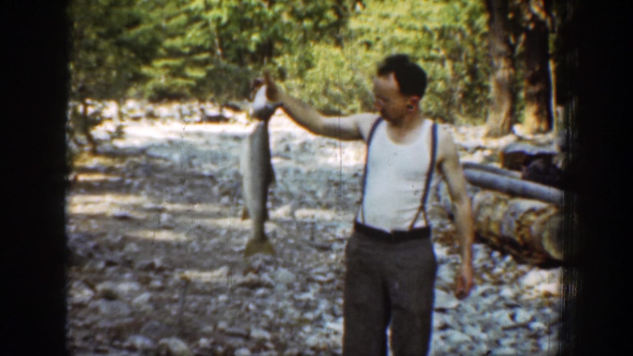 A man in suspenders lifts a large fish for the camera on a Chelan-area riverbank, with trees, rocks, and logs visible behind him.