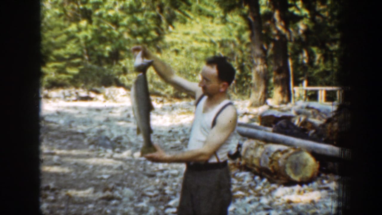 The 1939 Chelan home-movie frame shows the man presenting a large fish near a forested rocky stream with stacked logs behind him.