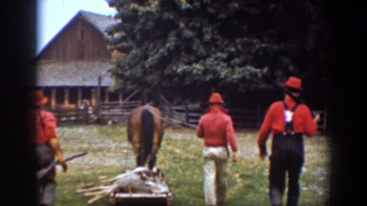 1939 Chelan, Washington frame with three men in red shirts, one carrying a rifle, following a horse-drawn wood sled toward a barn.