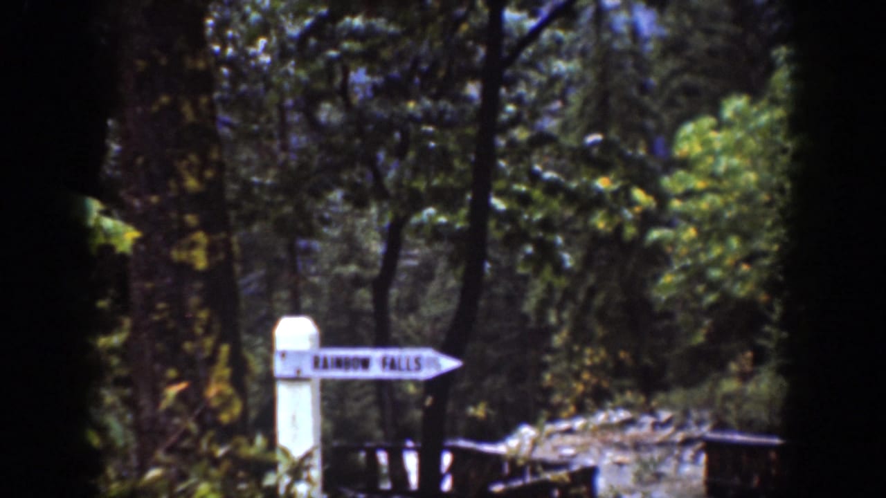 Home-movie frame of the Rainbow Falls trail sign amid trees, rocks, and greenery near Chelan, Washington.