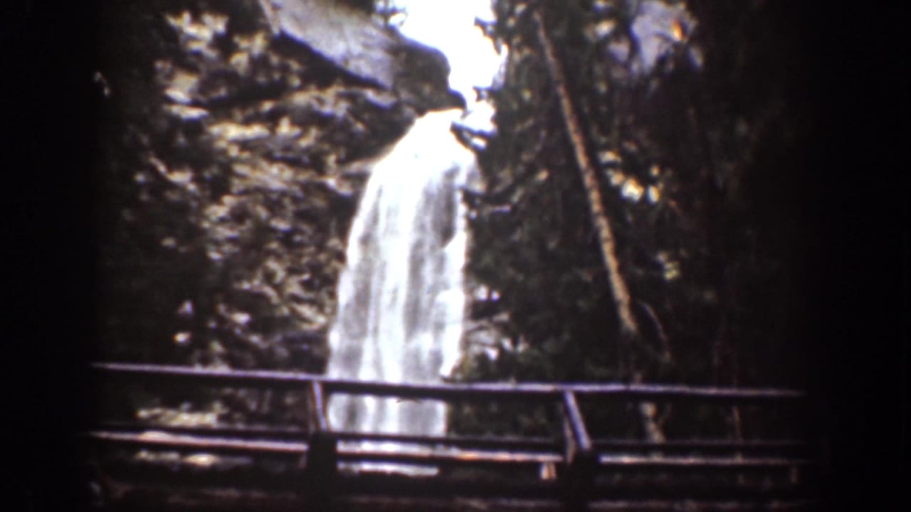 1939 Chelan, Washington home-movie frame showing a waterfall behind a narrow bridge in a dark rocky forest setting.