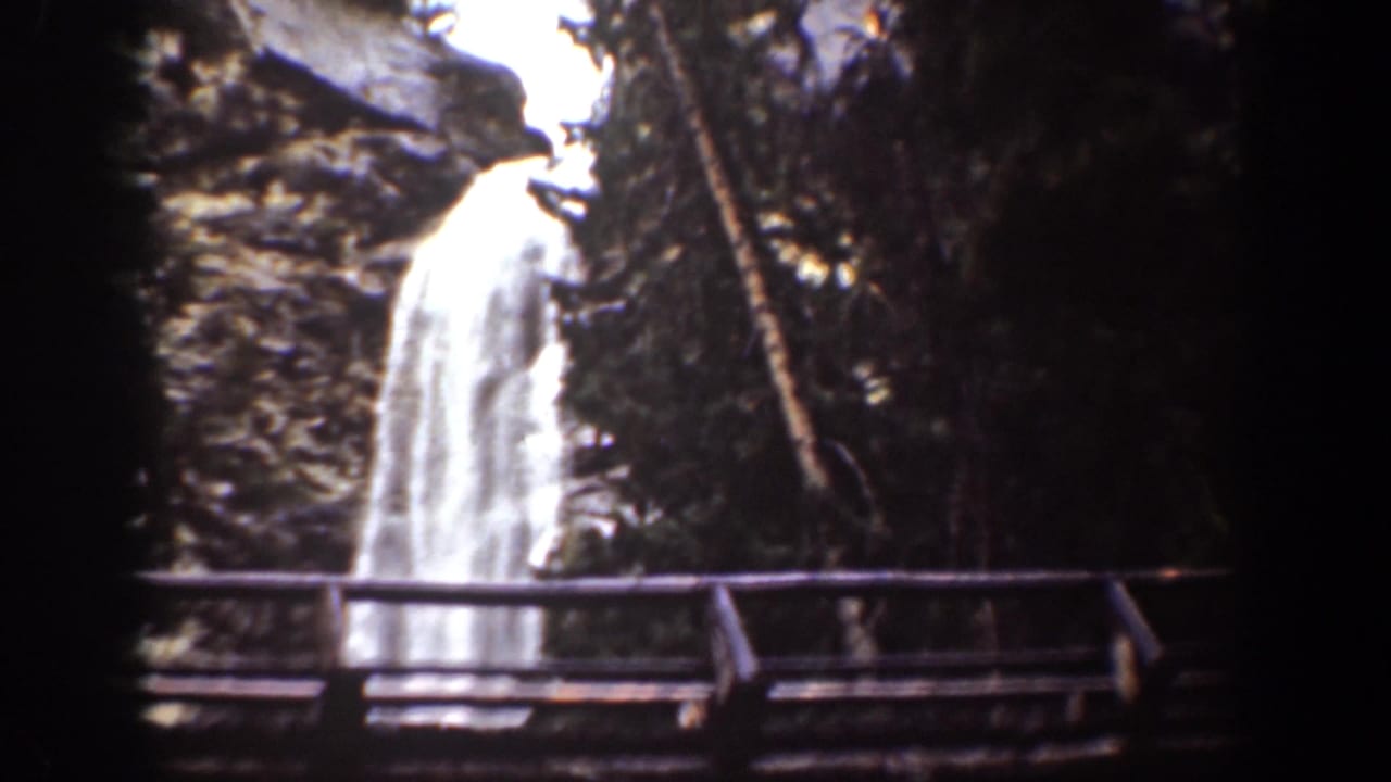 1939 Chelan, Washington view of a waterfall framed by trees, rock walls, and a wooden bridge railing.