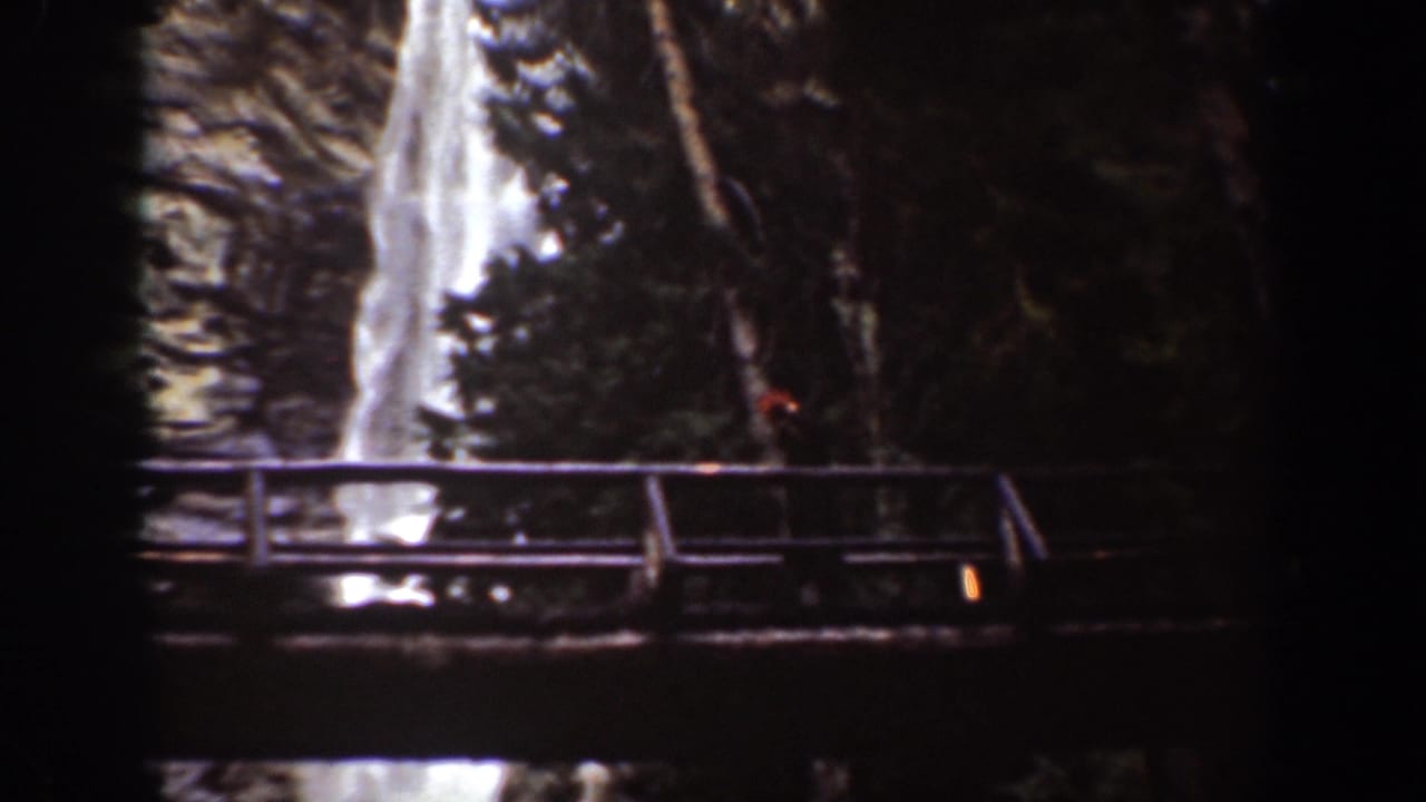 Chelan, Washington 1939 frame with a wooden bridge in the foreground and a waterfall dropping behind trees.