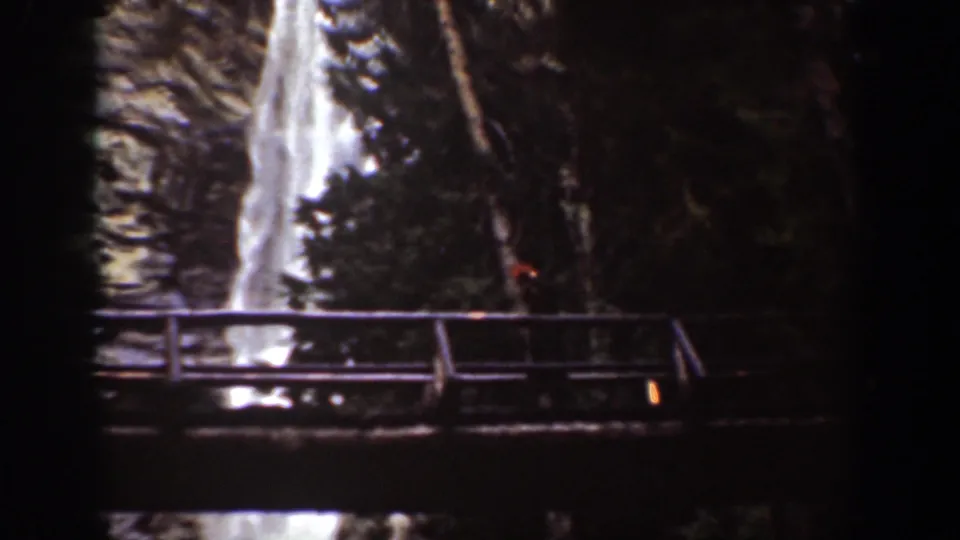 Chelan, Washington 1939 frame with a wooden bridge in the foreground and a waterfall dropping behind trees.
