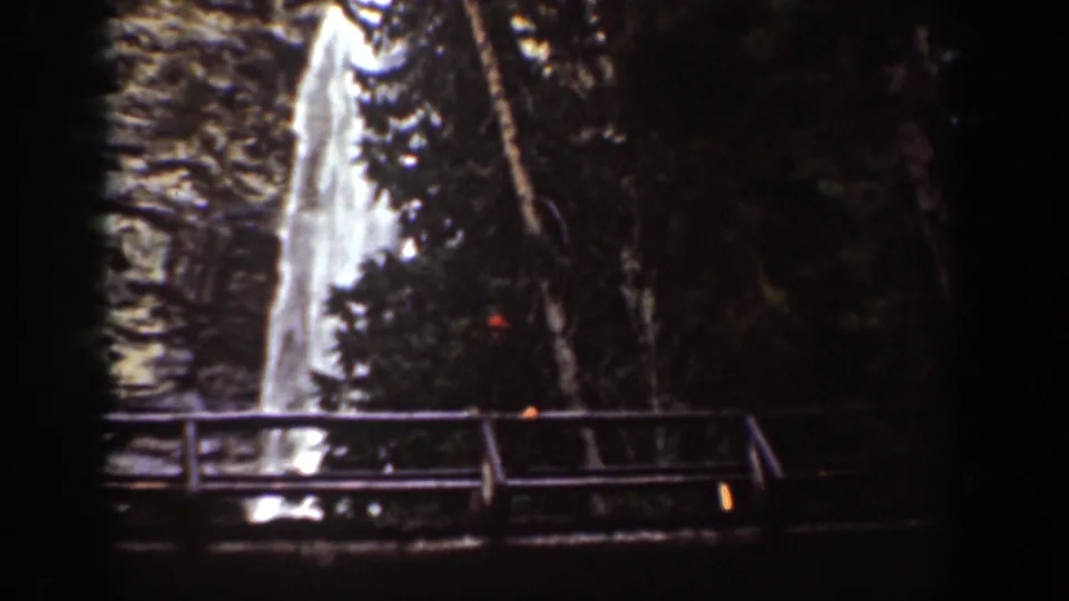1939 Chelan, Washington home-movie still of a waterfall beyond a wooden bridge and shaded evergreens.