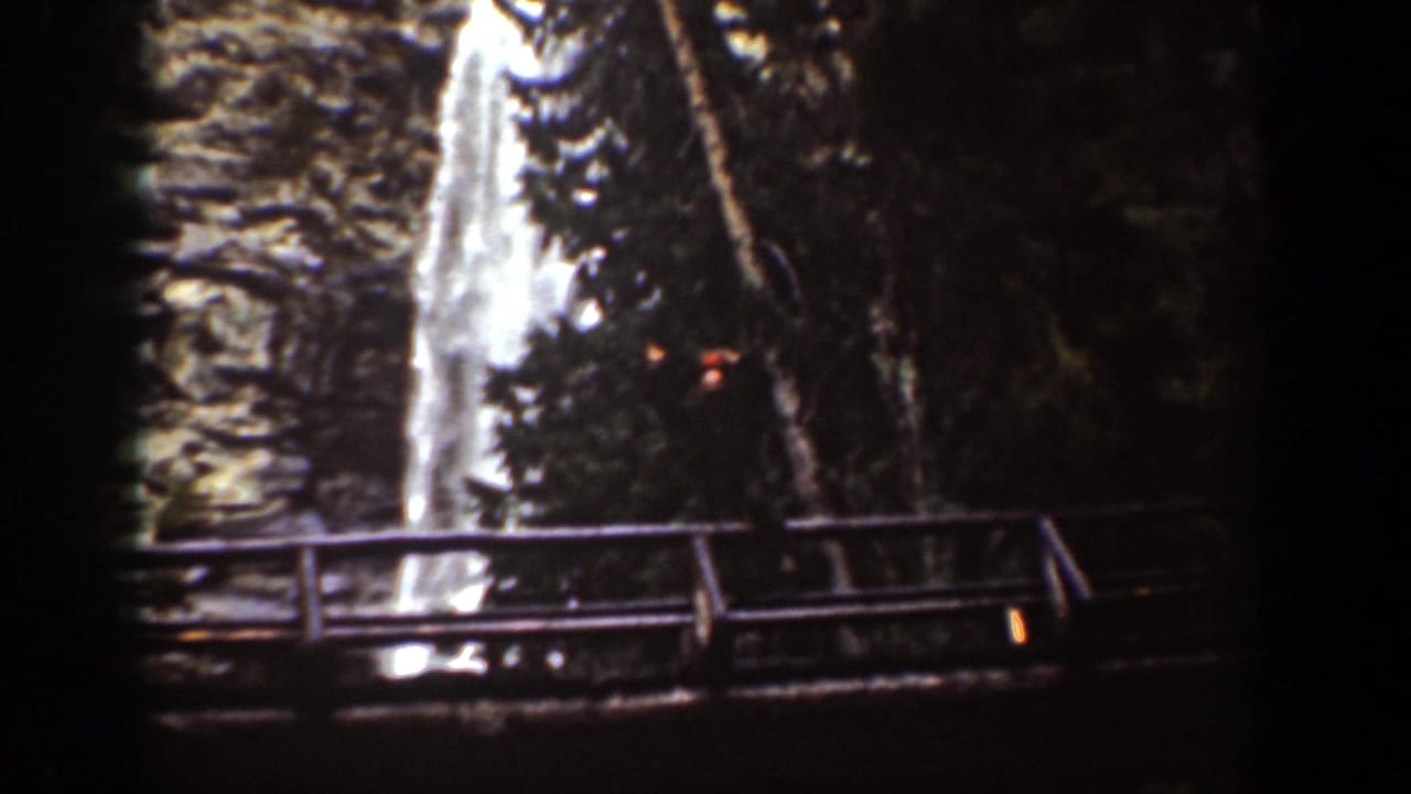 1939 Chelan, Washington frame with a small person on a wooden bridge before a tall waterfall in a forested gorge.