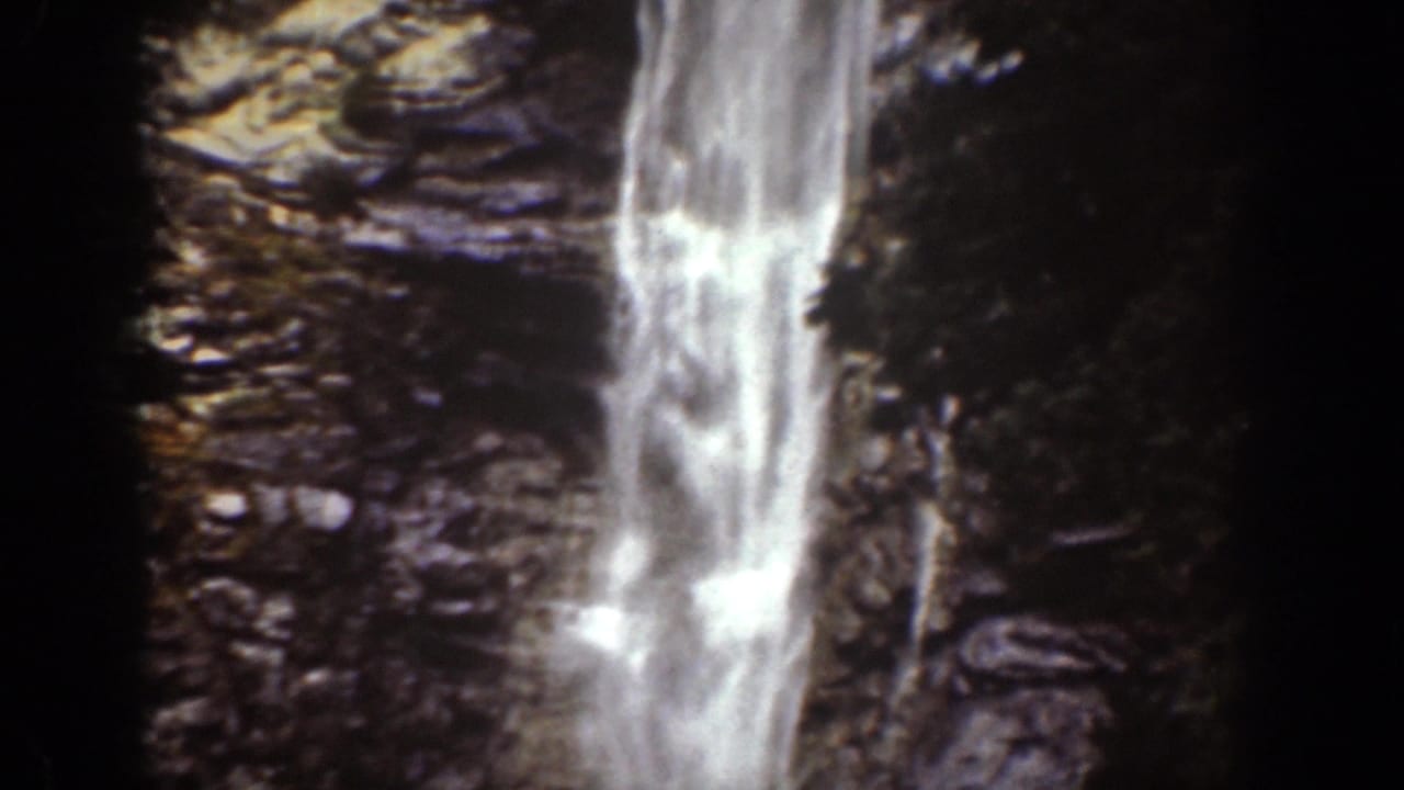 Waterfall dropping beside dark rock walls in a shadowed gorge.