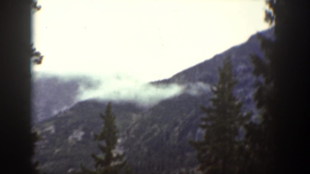 1939 Chelan-area mountain view with dark evergreen silhouettes, a sloping ridge, and low cloud or mist in the distance.