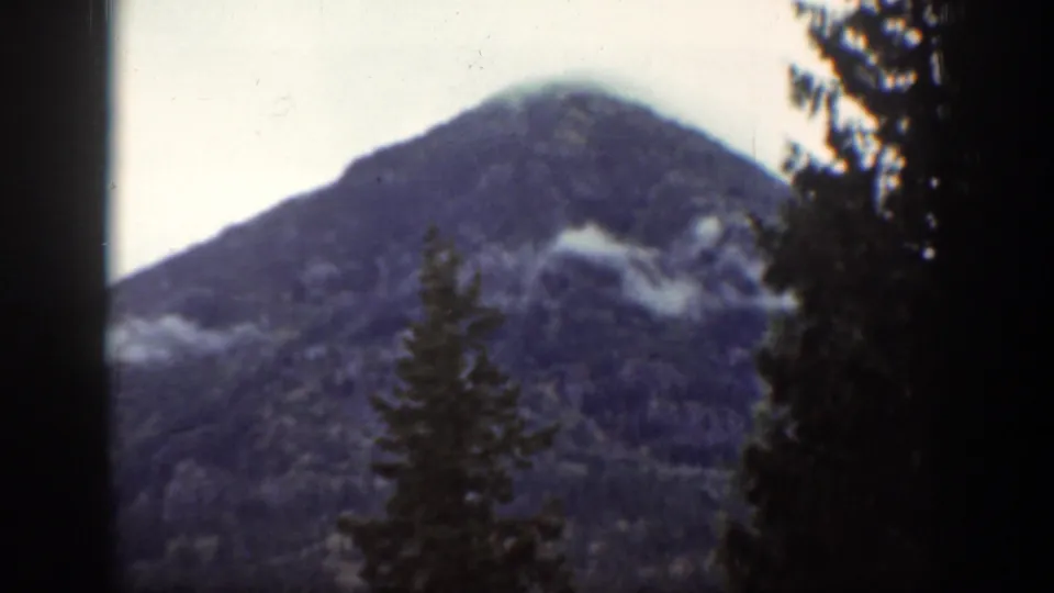 Chelan, Washington mountain forest scene with evergreen trees in front of a clouded peak, seen in faded 1939 8mm color.
