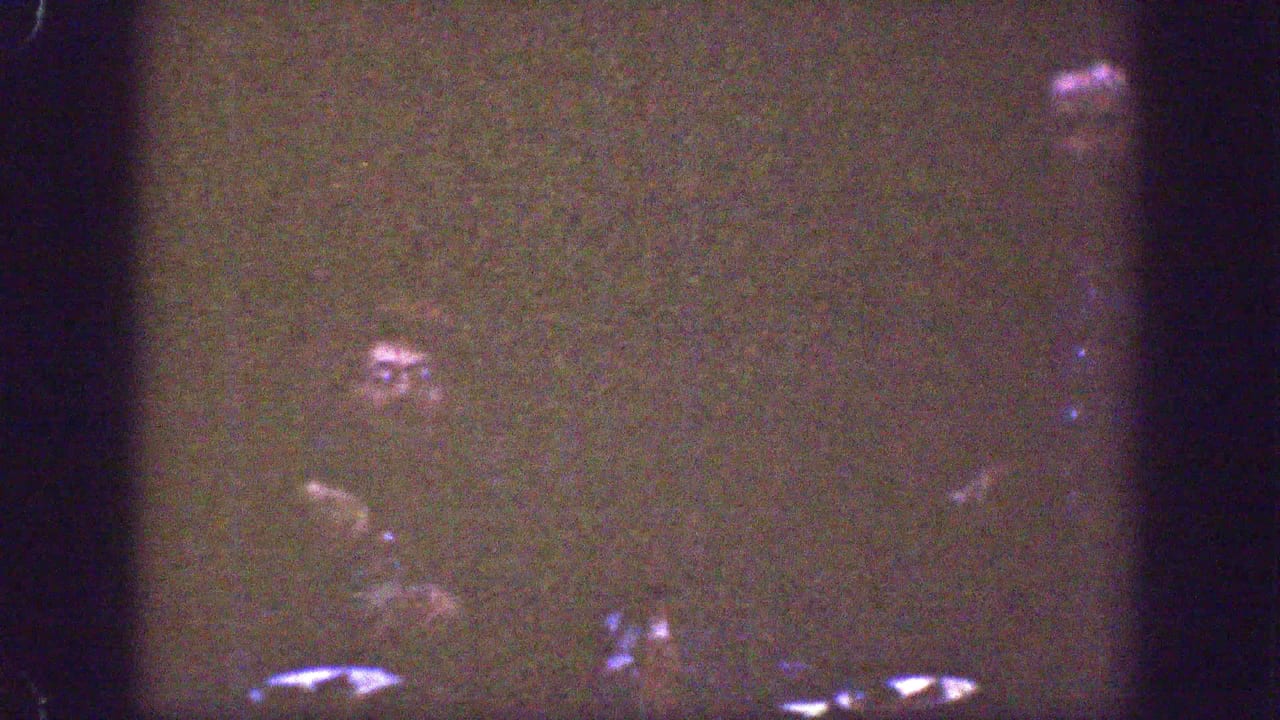 Grainy 1939 Chelan home-movie frame showing a dark gathering with partially lit adult faces and reflective table items.