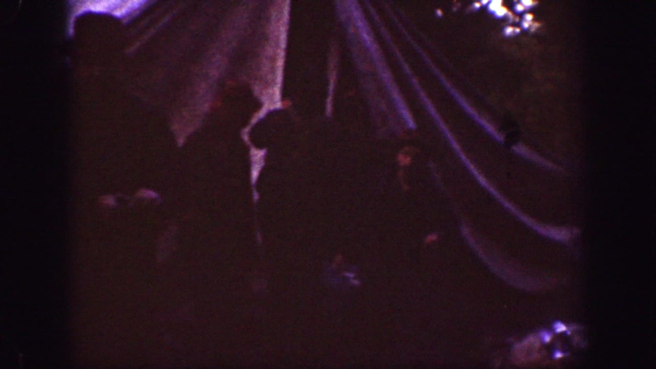 1939 Chelan campsite frame with a canvas tent or rainfly at right and a shadowed person standing nearby in low light.