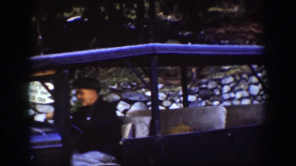 Chelan, Washington 1939 frame with a man in a dark hat seated inside an open-sided car near a stone wall and trees.