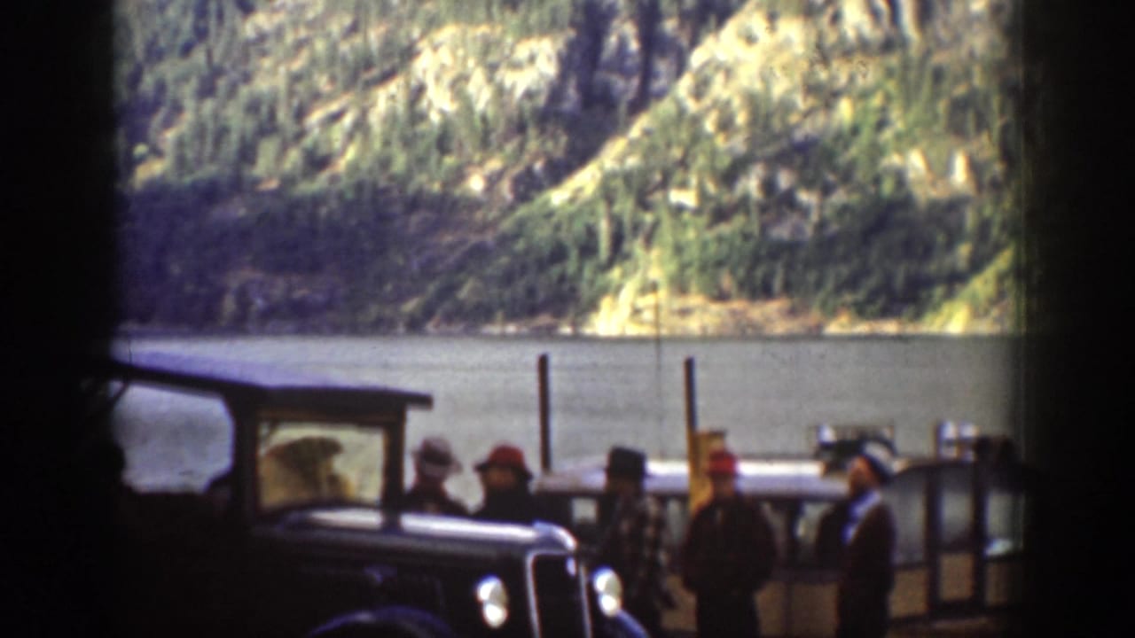 Chelan, Washington 1939 frame showing adults gathered near dark cars beside a lake with steep mountain slopes beyond.