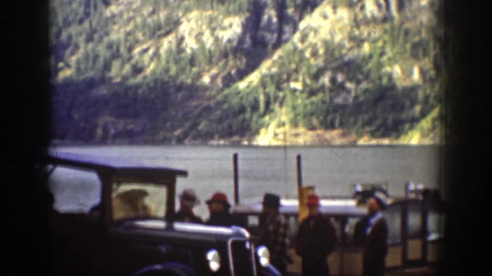Chelan, Washington 1939 frame showing adults gathered near dark cars beside a lake with steep mountain slopes beyond.