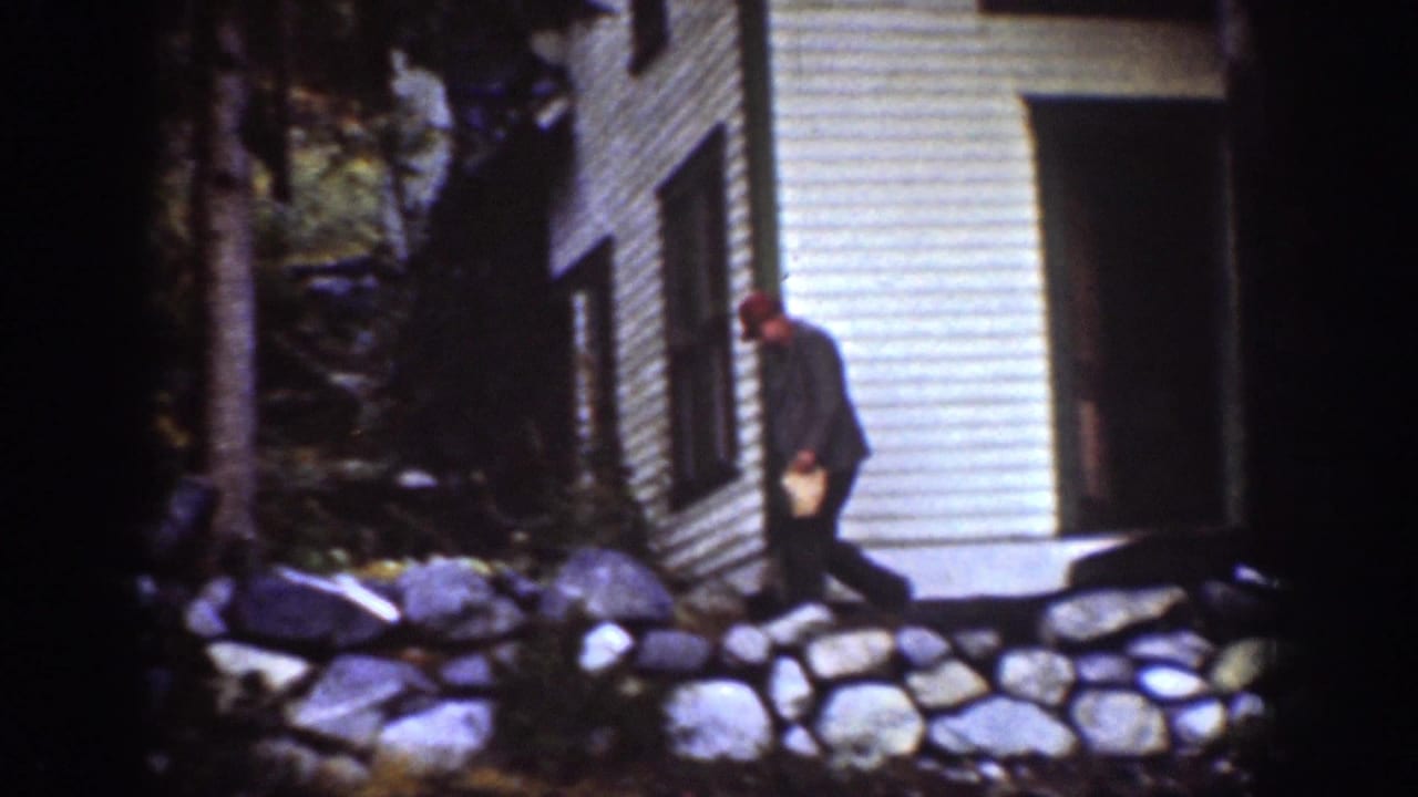 Man walking beside a white clapboard building and stone-lined path in a wooded area.