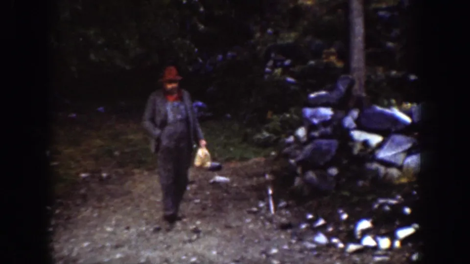 A man in a red hat and grey jacket walks through a wooded Chelan lakeside area carrying a small light-colored bag.