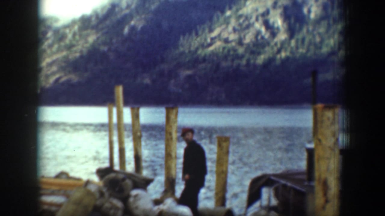 A man stands near wooden dock posts beside a lake in Chelan, with a forested hillside visible across the water.