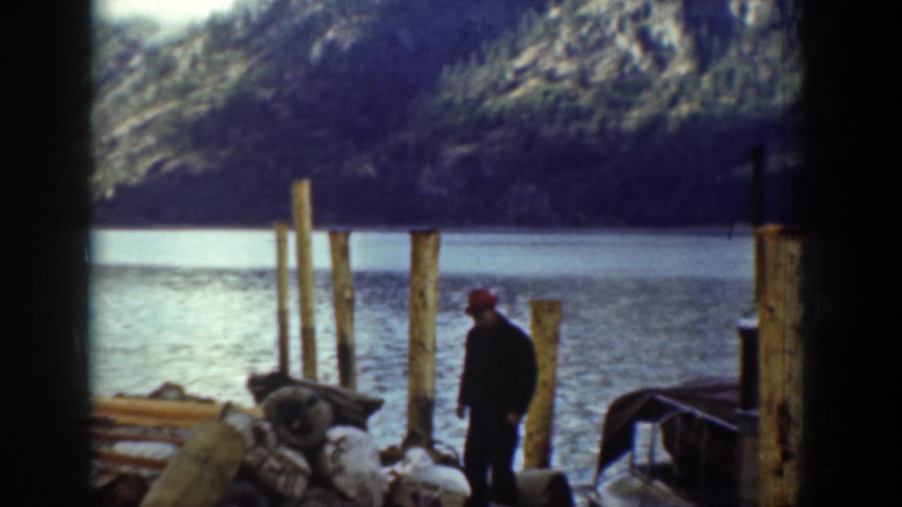 Man in a red cap stands near lakeside pilings with water and mountains behind him.