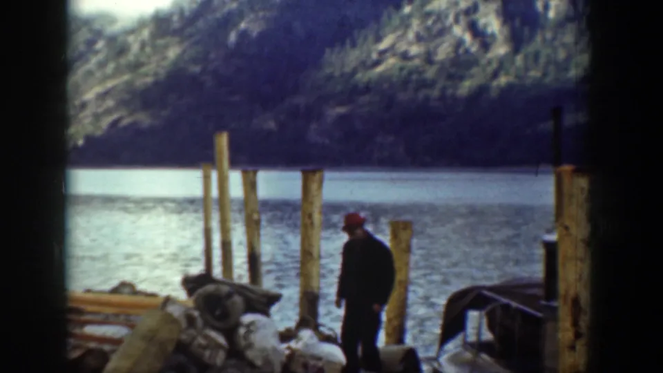In Chelan, Washington, 1939, a man in dark clothing and a red cap stands on a rocky shore near wooden pilings beside the lake.